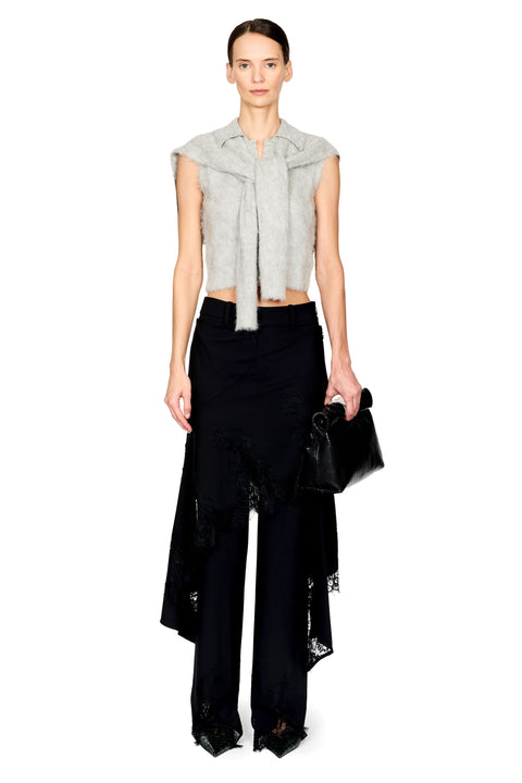 A model stands against a white studio background wearing a light gray sleeveless fuzzy cropped top with a tie detail at the collar, black wide-leg pants with layered ruffle and lace trim, and holding a black textured clutch.