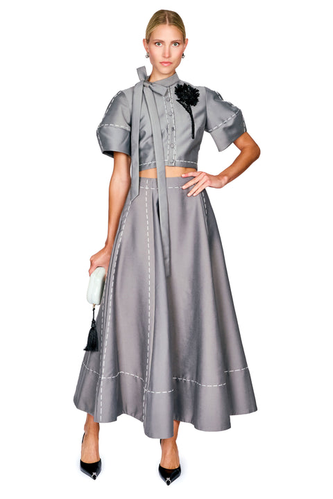 A model wearing a gray two-piece set consisting of a short-sleeve cropped top with a tie-neck detail and a matching long skirt, styled with black heels and a small black and white handbag against a white background.