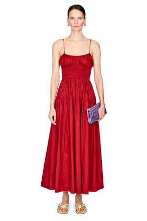 A woman modeling a long red spaghetti-strap dress with a ruched bodice and a flared skirt, holding a blue and red striped clutch against a plain white background.