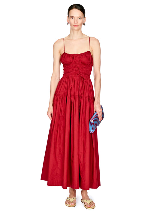 A woman modeling a red spaghetti-strap maxi dress with a smocked bodice and tiered skirt, holding a blue striped clutch against a plain white background.