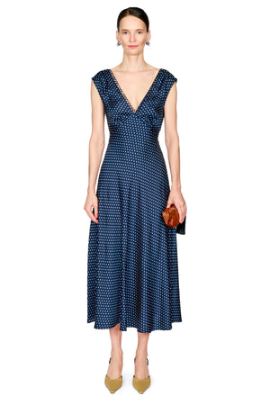 A woman modeling a navy blue midi dress with a white polka dot pattern, featuring a V-neckline and cap sleeves, paired with tan pointed-toe heels and holding a small brown clutch against a plain white background.