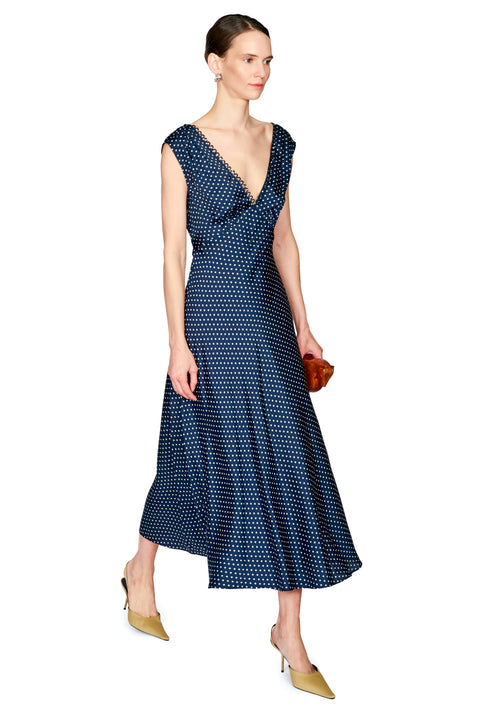 A woman modeling a navy blue midi dress with a white geometric pattern, featuring a deep V-neckline and cap sleeves, paired with tan pointed-toe heels and holding a small brown clutch against a plain white background.