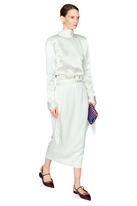 A model wearing a white satin long-sleeve mock-neck dress with a matching belt, holding a striped clutch, and wearing brown pointed-toe flats against a white background.