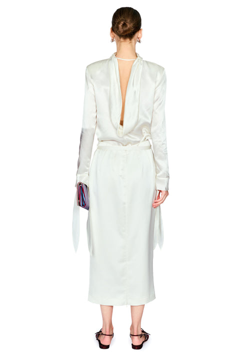A model shown from the back wearing a white satin long-sleeve dress with a deep V-back, tie waist, and long ribbon tails, paired with black strappy heels.