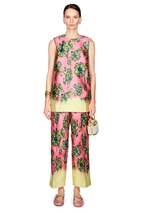 A model stands against a white background wearing a sleeveless pink top with a green and blue floral print and a yellow-green hem, paired with matching wide-leg pants, a gold cuff , and a small woven handbag.