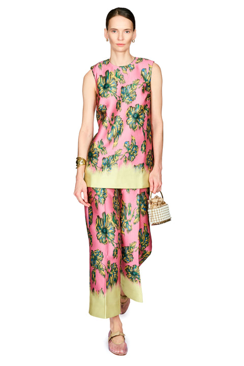 A model wearing a sleeveless pink two-piece outfit with a green and teal floral print, featuring a top and matching wide-leg pants with a solid light green hem. She is holding a small woven handbag and wearing gold jewelry against a plain white background.