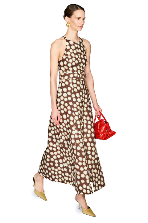 A woman modeling a sleeveless halter-neck maxi dress with a brown and beige polka dot pattern, paired with gold pointed-toe heels and holding a small red handbag against a plain white background.
