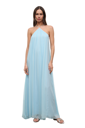 A woman modeling a light blue halter-neck maxi dress with a flowing, pleated chiffon-like fabric, paired with silver sandals, against a plain white background.