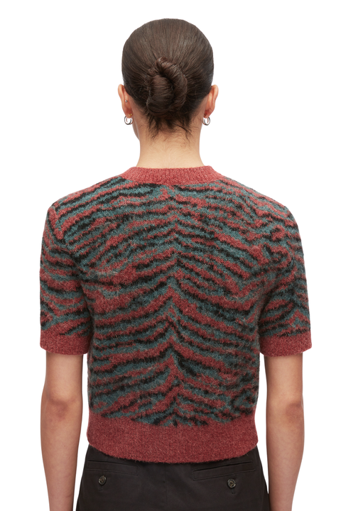 Back view of a model wearing a short-sleeve knit sweater with a textured chevron pattern in rust red and dark teal, paired with black trousers against a plain white background.