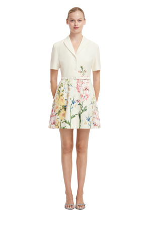 A model wearing a short-sleeve cream-colored wrap-style mini dress with a colorful floral print on the skirt, paired with clear high-heeled sandals, against a plain white background.