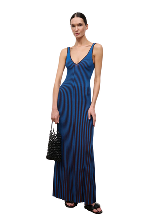 A model wearing a sleeveless, deep V-neck maxi dress with vertical blue and brown stripes, holding a black woven handbag against a solid white background.