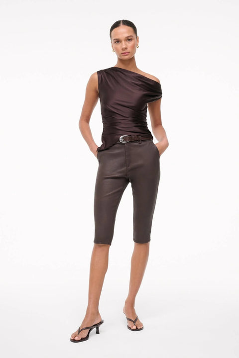 Woman wearing an asymmetrical, brown silk top that features an off the shoulder design and draped ruching throughout, paired with brown capris and black sandals. 