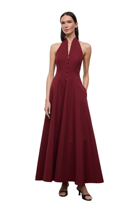 A woman modeling a sleeveless, halter-neck, floor-length maxi dress in deep burgundy. The dress features a buttoned bodice and a flowing skirt, styled with black heeled sandals against a plain white background.