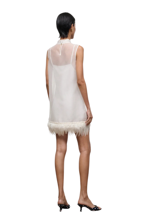 A woman shown from the back wearing a sleeveless white tulle dress with a feathered hem and a high neck, paired with black heeled sandals, against a plain white background.