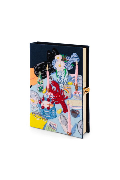 A book clutch featuring a colorful, whimsical illustration of a dinner table scene with a lobster, flowers, candles, and a cat, secured with a gold-toned band on a white background.