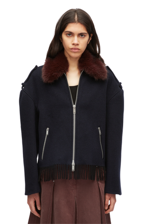 A model wearing a dark navy blue jacket with a large dark brown fur collar, silver zippers, and fringe trim at the hem, paired with a brown skirt against a plain white background.