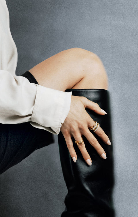 A woman holding her hand over her black boots and wearing a Spinelli Kilcollin ring that features a thin 18k yellow gold ring with pave white diamonds, a thick 18k yellow gold band, another thing 18k yellow gold band with pave white diamonds, and another thick 18k yellow gold band, all connected with three 18k yellow gold connector rings on her index finger and another Spinelli ring on her ring finger. 