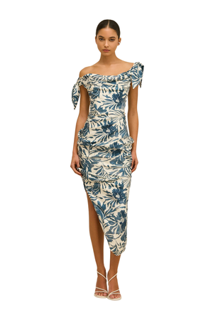 A woman modeling a fitted off-the-shoulder midi dress with a blue and cream tropical floral print, paired with white strappy high-heeled sandals, shot against a solid white background.