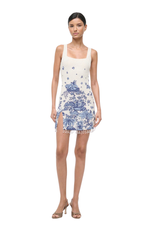 A model wearing a sleeveless white mini dress with a blue traditional landscape print and fringe trim, paired with nude high-heeled sandals, against a plain white background.