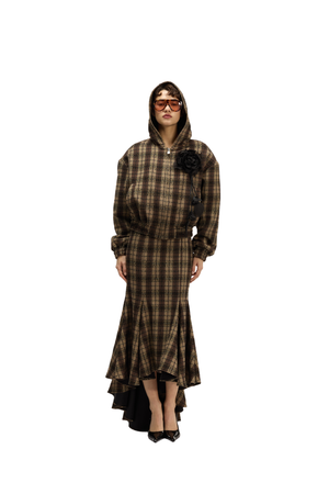 A model wearing a brown and black plaid hooded dress with a high-low hem, a black rose embellishment on the chest, and black pointed shoes, set against a plain white background.