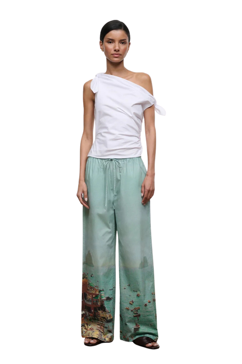 A model wearing a white asymmetrical one-shoulder top and wide-leg mint green pants with a scenic landscape print near the hem, standing against a plain white background.