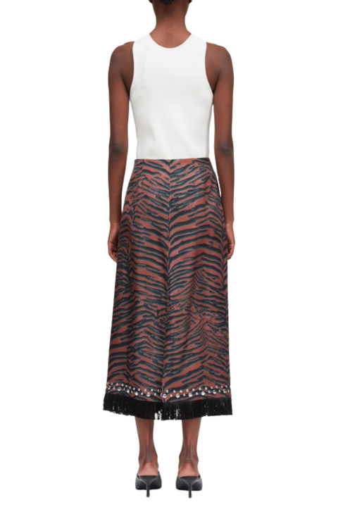 A model shown from the back wearing a sleeveless white top and a high-waisted midi skirt with a zebra-like pattern in rust red, black, and dark gray, finished with a decorative embroidered hem and black fringe.