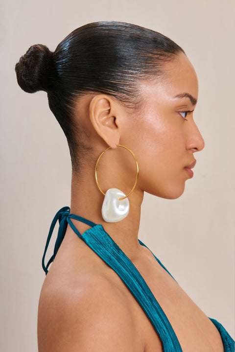 Side angle of a woman wearing thin gold hoop earrings that feature abstract, asymmetrical pearl charms. 