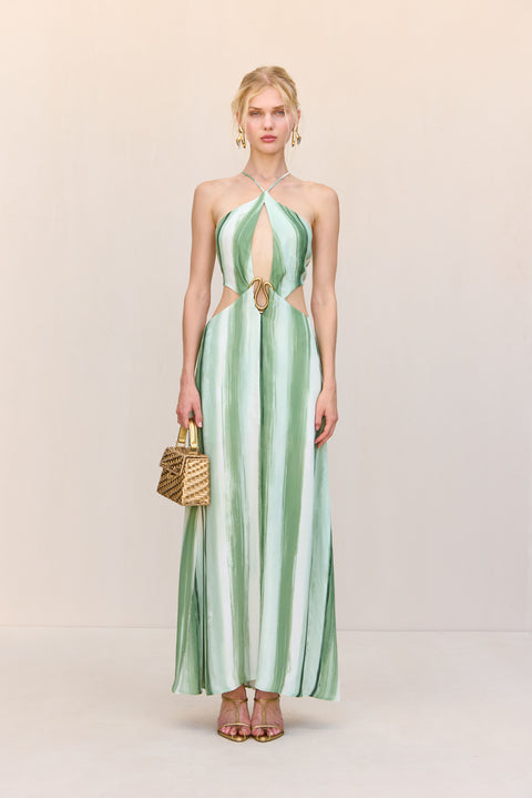 Woman wearing a green and white painted stripe maxi dress that features a halter neck and large cutouts at the chest and waist with gold-toned abstract hardware at the center, paired with gold heels and a handbag. 