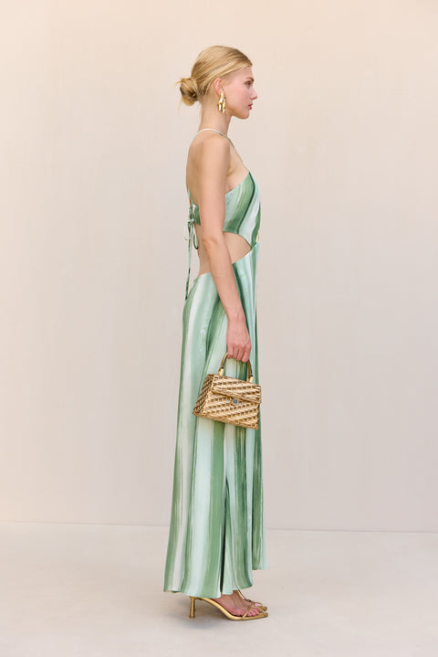 Side view of a woman wearing a green and white painted stripe maxi dress that features a halter neck and large cutouts at the chest that tie in the back, paired with gold heels and a handbag. 