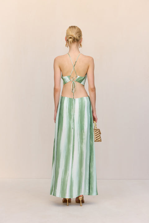 Back view of a woman wearing a green and white painted maxi dress that features n open back and spaghetti straps that cross at the shoulders and tie in a lace up design in the back, paired with gold heels and handbag.