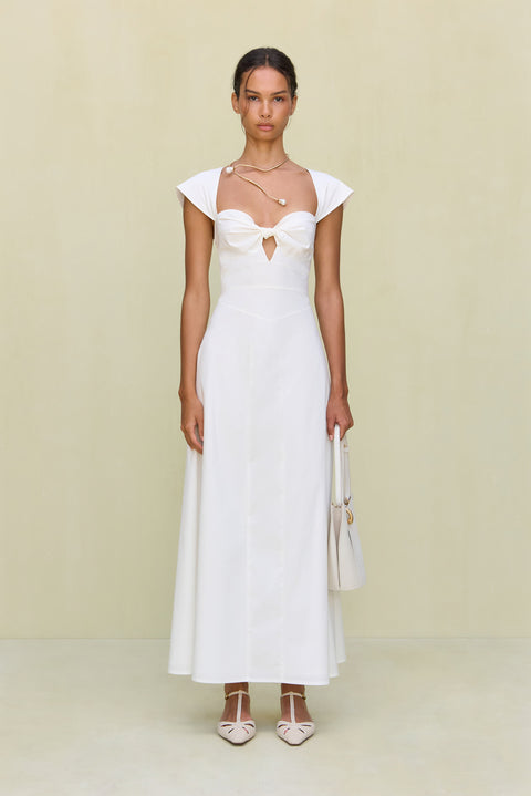 Woman wearing a white maxi poplin dress that features a sweetheart neck, cap sleeves, and a cutout at the bust, paired with white sandals. 