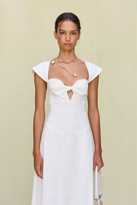 Close up of a woman wearing a white maxi poplin dress that features a sweetheart neck, cap sleeves, and a cutout at the bust, paired with white sandals. 