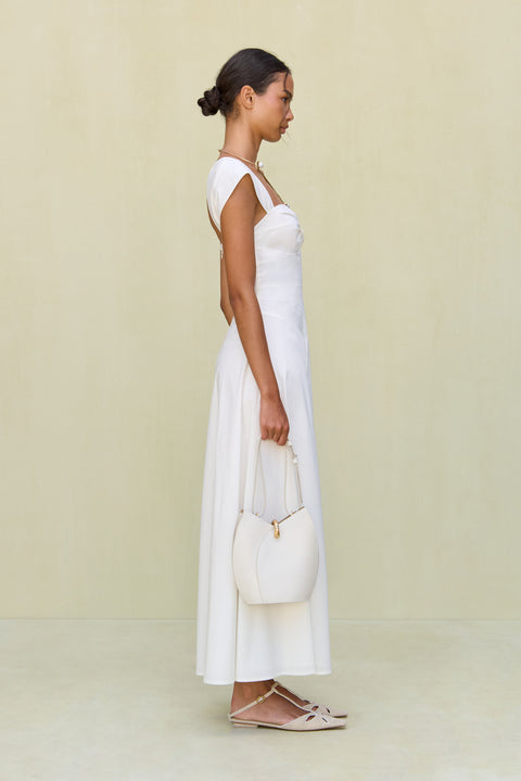 Side view of a woman wearing a white maxi poplin dress that features a sweetheart neck, cap sleeves, and a cutout at the bust, paired with white sandals. 