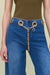 Detail shot of blue low-rise, wide leg jeans that feature gold metal grommets along the waist and a Cult Gaia logo chain belt, paired with a green bodysuit. 