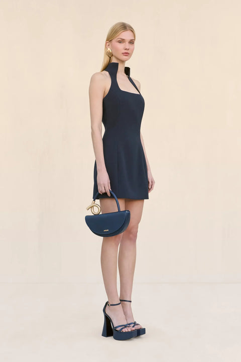 Side view of a woman wearing a navy sleeveless mini dress that features a stand collar and a square neck with a tailored fit, paired with navy shoes and handbag. 