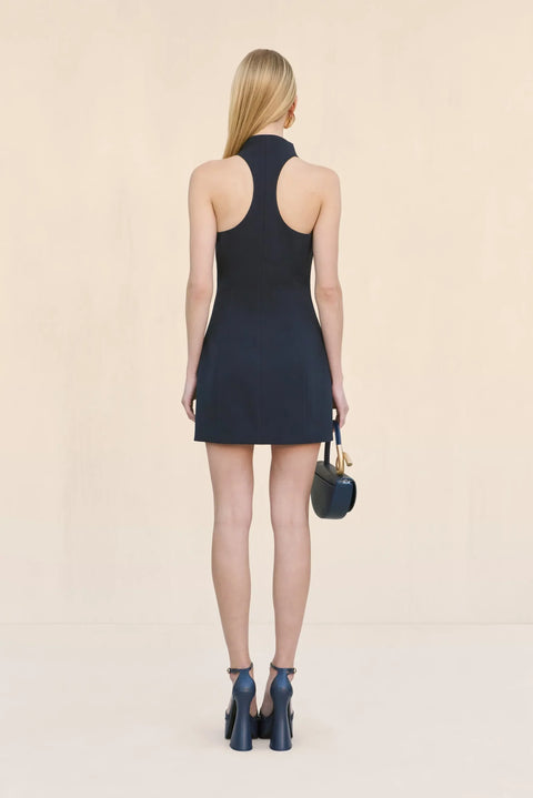 Back view of a woman wearing a navy sleeveless mini dress that features a stand collar, racerback detail, and a tailored fit, paired with navy heels and a navy handbag. 