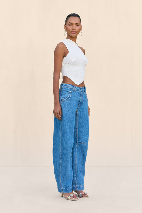 Side-facing woman wearing a white knit off the shoulder crop top that features a cutout at the rib cage and an asymmetrical hem, paired with blue wide leg jeans.