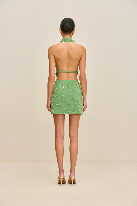 Back view of a women wear a green jersey mini dress that features a halter neck and open back with a strap across the midsection, adorned with droplet shaped, iridescent glass beads, paired with gold heels.