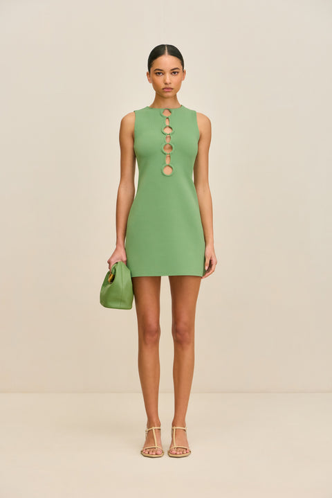 Woman wearing a sleeveless, green knit mini dress that features a high neck and four ring cutouts at the chest, paired with a gold handbag and gold heels. 