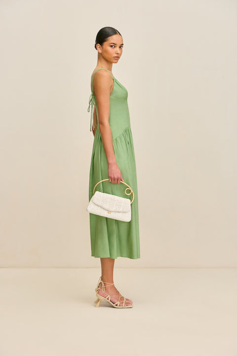 Side view of a woman wearing a green, basque waist midi dress that features a double spaghetti strap detail, a deep-V and a small keyhole cutout at the bust, paired with tan heels and a white handbag. 