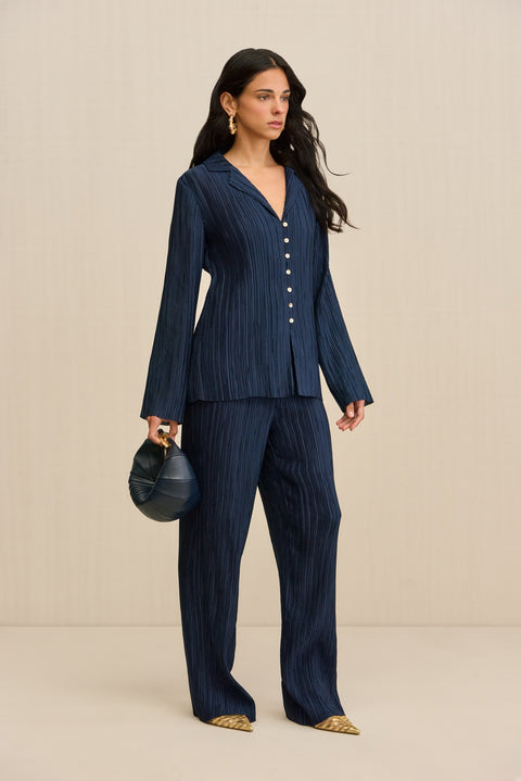 Side-facing woman wearing a navy plisse button down shirt that features pearlescent contrast buttons, paired with matching pants and a navy handbag. 