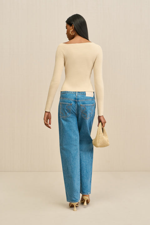 Back-facing woman wearing a cream long sleeve shirt with a ring detail at the shoulder, paired with blue jeans. 