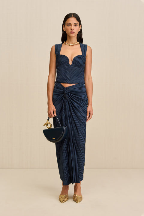 Woman wearing a navy blue midi skirt that features an asymmetrical waistline with knotted ruching at the hip, paired with a matching top and gold heels. 
