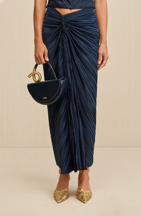 Close up of a woman wearing a navy, ankle-length midi skirt that features an asymmetrical waistline with knotted ruching at the hip, paired with gold heels and a black handbag. 