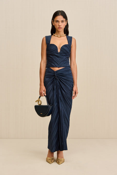 Woman wearing a navy crop top crafted from plisse fabric, featuring a plunging sweetheart neckline and corset style seaming, paired with a matching skirt and gold heels. 