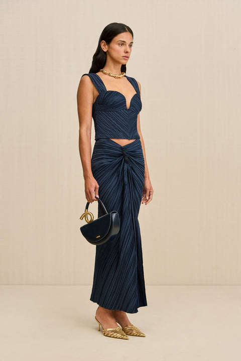Side-facing woman wearing a navy crop top crafted from plisse fabric, featuring a plunging sweetheart neckline and corset style seaming, paired with a matching skirt and gold heels. 