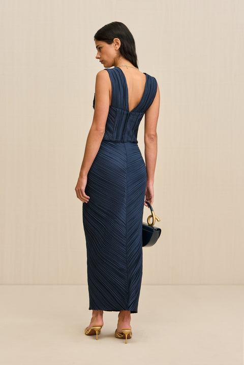 Back-facing woman wearing a navy blue midi skirt that features an asymmetrical waistline with knotted ruching at the hip, paired with a matching top and gold heels. 
