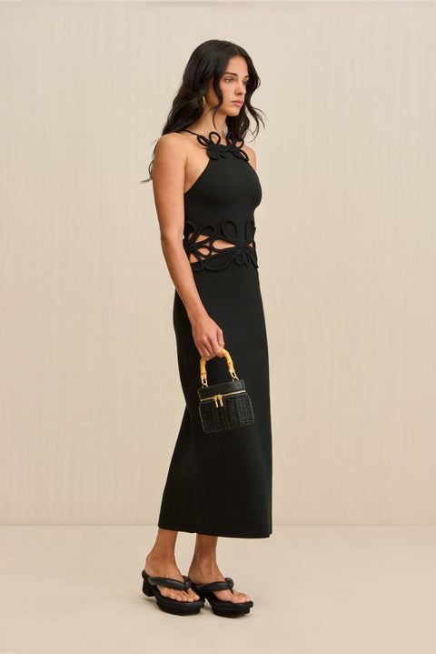 Side-facing woman wearing a black midi dress that features waist cutouts and floral piping overlays at the waist and neck. 