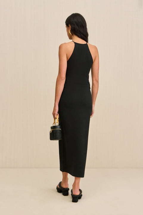 back-facing woman wearing a black midi dress that features waist cutouts and floral piping overlays at the waist and neck. 