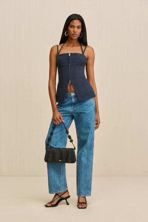Woman wearing a navy tank top that features a double strap detail, flattering seaming, and a double front zipper, paired with jeans and black heels. 
